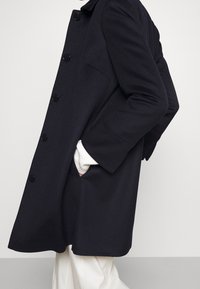 Navy coat made of wool blend, featuring large black buttons, a relaxed fit, side pockets, and a collar. Accents with a white knit cuff visible.