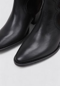 Botas de cuero negro hasta el tobillo con punta afilada y textura suave, que presentan costuras decorativas y un bajo tacón en bloque.