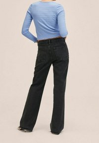 Blaues, langärmliges, kurz geschnittenes Strickoberteil und schwarze weite Jeans mit Rückentaschen und fransigem Saum. Modell steht mit dem Rücken zu einem neutralen Hintergrund.