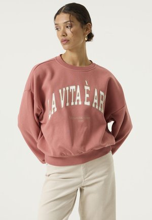 Vrouw staat met de handen achter haar rug, draagt een roze sweatshirt met de tekst "LA VITA È ARTE" en een crèmekleurige broek, kijkt opzij.