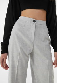 Kobieta nosząca jasnoszare, w kratkę spodnie z wysokim stanem, z czarnym guzikiem, oraz krótki czarny, prążkowany sweter, widoczna od połowy brzucha w dół.