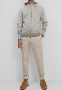 Sudadera con cremallera en gris claro, con un tejido texturizado y capucha ajustable, combinada con pantalones cargo beige y zapatos marrones con cordones.