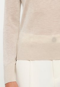 Pull en tricot beige clair avec un ourlet à côtes et des poignets côtelés, offrant une texture lisse et une légère variation de couleur dans le tissu.