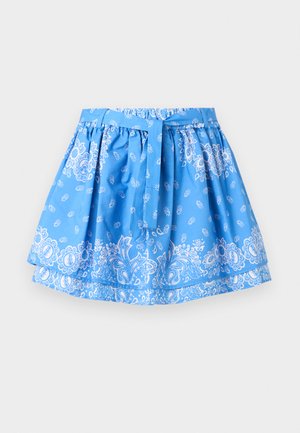 Młoda kobieta stoi, ubrana w biały, bezrękawnikowy crop top, niebieską wzorzystą spódnicę zapinaną na kokardę oraz czarne sandały na obcasie na jednolitym tle.