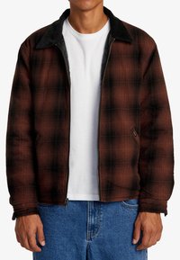 Brun och svart rutigt zip-up jacka med en kordfloderkant, texturerad tyg och sidofickor, bärs över en vit skjorta och denimbyxor.