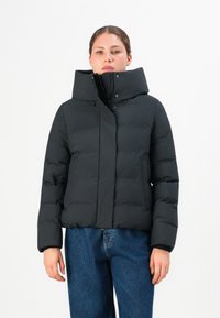 Femme portant une veste rembourrée noire avec un col montant et une fermeture éclair, associée à un jean bleu, se tenant devant un fond blanc uni.