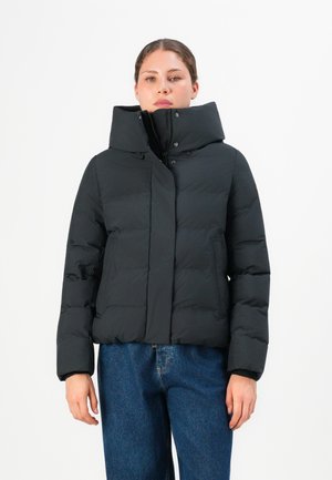 Vrouw in een zwarte pufferjack met hoge kraag en rits, gecombineerd met blauwe jeans, staand tegen een egale witte achtergrond.
