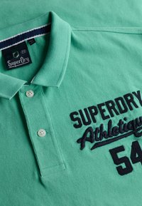 Polo vert en tissu texturé, présentant un design à col, une patte de boutonnage à deux boutons, et un logo brodé "SuperDry Athletic" en marine.