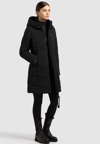 Schwarze Daunenjacke mit Kapuze, mit einem gesteppten Design, Reißverschluss und Daumenlöchern. Getragen mit schwarzen Leggings und Ankle Boots.