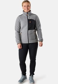Grijze fleecejas met zwarte accenten, ritszakken en hoge kraag. Gecombineerd met zwarte pantalons met ritszakken en grijze sneakers.