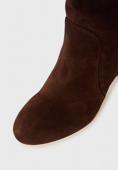 Primo piano di un stivaletto alla caviglia in suede marrone scuro con punta arrotondata e texture liscia su uno sfondo bianco.