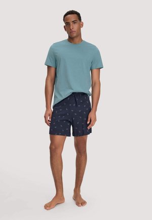 Junger Mann steht barfuß, trägt ein türkisfarbenes Kurzarm-T-Shirt und marineblaue Shorts mit Paisley-Muster auf einem schlichten hellen Hintergrund.