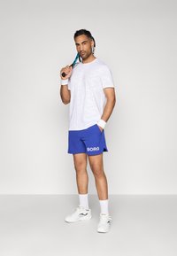 Λευκό T-shirt με καμουφλάζ, μπλε σορτσάκι με κείμενο "BORG", λευκά βραχιόλια, λευκές κάλτσες και αθλητικά παπούτσια. Κρατάει μια ρακέτα τένις.