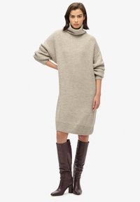 Vestido suéter de punto oversized en beige claro con cuello alto, puños acanalados y dobladillo. Se lleva con botas ajustadas hasta la rodilla de color púrpura profundo.