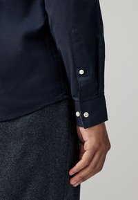 Camicia blu navy con bottoni, colletto strutturato e polsini, con bottoni bianchi e tessuto dalla texture liscia.