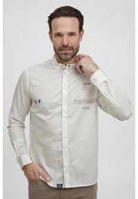 Világos bézs gombos ing, hímzett szöveggel és logókkal, hosszú ujjakkal, gallérral és diszkrét textúrájú anyaggal.