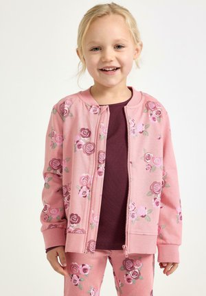 Leende ung flicka som bär en rosa blommig zip-up jacka och matchande byxor med en mörklila tröja under, stående mot en vit bakgrund.