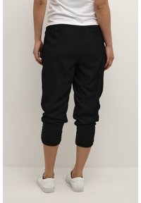 Pantalons capri noirs avec une coupe ample, design de chevilles froncées et taille élastique, associés à des baskets blanches. Texture lisse.