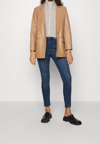 Tan läderblazer, grå ribbad tröja med hög hals, mörkblå skinny jeans och svarta slip-on skor. Enkelt design, slät textur, diskreta accenter.