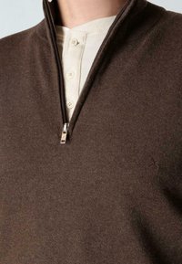 Lähikuva henkilöstä, joka pukeutuu ruskeaan vetoketjulliseen neuleeseen, jonka alla on vaalea vohvelikuvioinen henley-paita, jossa kolme nappia näkyvисс.