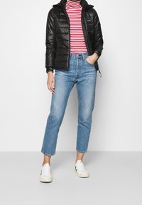 Sort pufferjakke med quiltet design, rød og hvid stribet rullekrave, lys blå højhævede jeans og hvide sneakers.