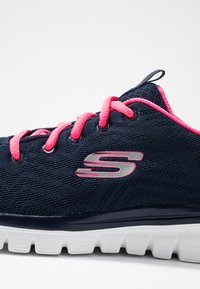 Strømpesko i slankt marineblått med rosa lisser, mesh-overdel og metallisk logo. Har en polstret hvit såle med teksturert grep.