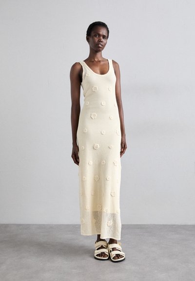 Robe sans manches en tricot jaune pâle avec des appliqués floraux. Longue jusqu'au sol, en tissu texturé, avec décolleté en forme de scoop et dos ouvert.