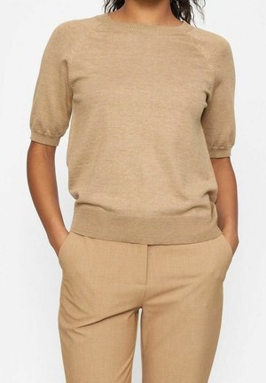 Nainen, joka pukeutunut beigeen lyhythihaiseen neulepaitaan ja siihen sopiviin beigeihin housuihin, kädet taskuissa, taustalla yksinkertainen valkoinen tausta.