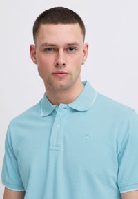 Vaaleansininen polo-paita, jossa on tekstuurinen kangas, kauluksessa kaksi nappia ja hienovarainen logo vasemmalla rinnassa. Valkoiset yksityiskohdat kauluksessa.