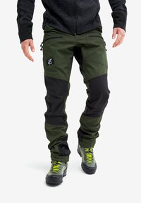 Legs wearing green and black outdoor pants with zip pockets and a black patch, paired with gray hiking boots with yellow laces.