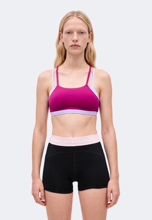 Une femme porte un soutien-gorge de sport magenta à bretelles lilas pâle et un short noir ajusté sur un fond clair uni.
