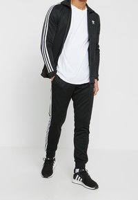 Svart atletisk träningssuit med vita ränder, bestående av en zip-up jacka och slim-fit byxor. Bärs med en vit t-shirt och svarta sneakers.