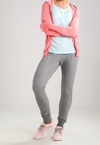 Person in einem korallefarbenen Zip-Up-Hoodie, einem hellblauen Shirt mit Text, grauen Jogginghosen und rosa Sneakers, steht mit verschränkten Armen.