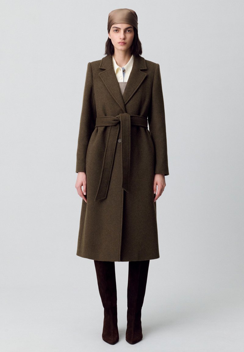Cappotto lungo in lana verde oliva con vita cinturata, ampi rever e chiusura a bottoni. Indossato su una camicia chiara e abbinato a stivali marroni.