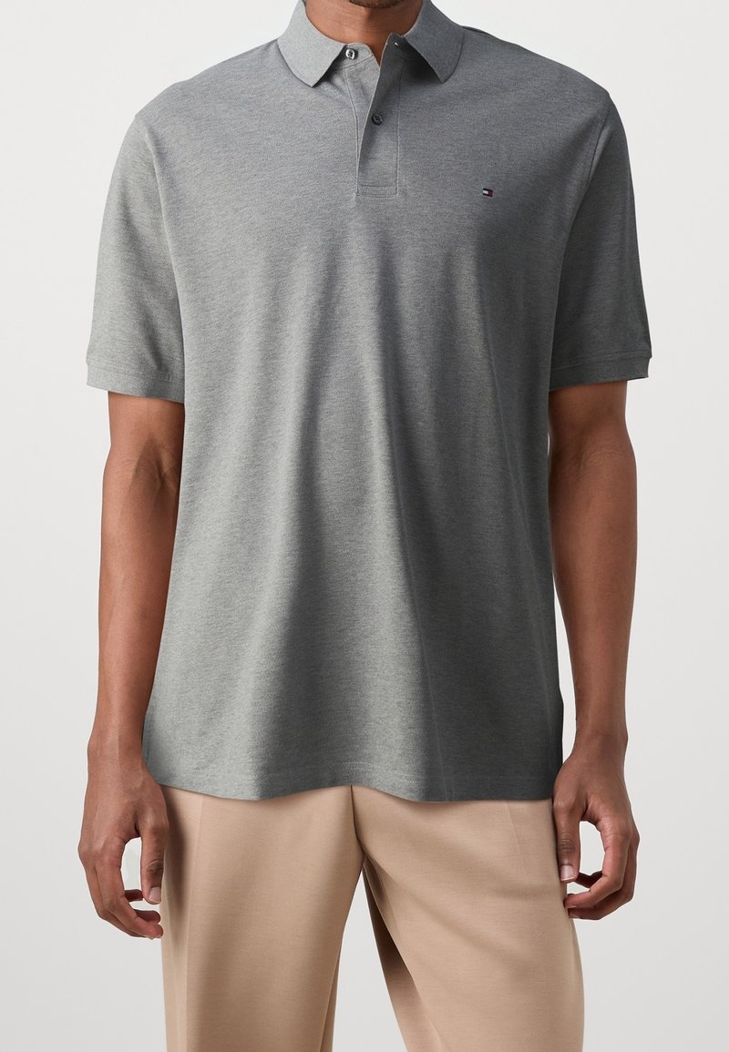 Homme portant un polo gris à manches courtes et un pantalon beige, debout, les bras relâchés le long du corps, devant un fond uni.