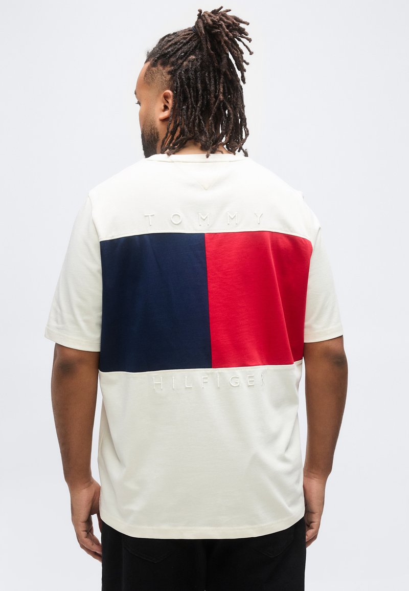 Άνδρας με ντραντλοκς που φοράει κρεμ μπλουζάκι με μεγάλα κόκκινα και ναυτικά μπλοκ χρωμάτων και ανάγλυφο κείμενο «Tommy Hilfiger» στην πλάτη.