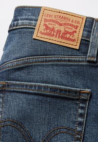 Jeans de denim azul com um remendo de couro castanho apresentando "LEVI STRAUSS & CO." e um design de rebites. Acentos de costura e dois bolsos traseiros visíveis.