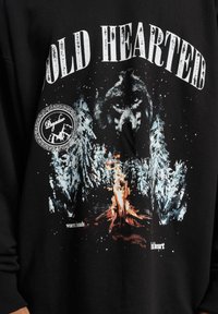 Melnā sporta jakā ir attēlots vilks un priedes, ar dzīvīgām krāsām attēlota ugunskura ainava. Uz tās ir teksts "AUKSTĀ SRDSTS" un "siltas rokas".