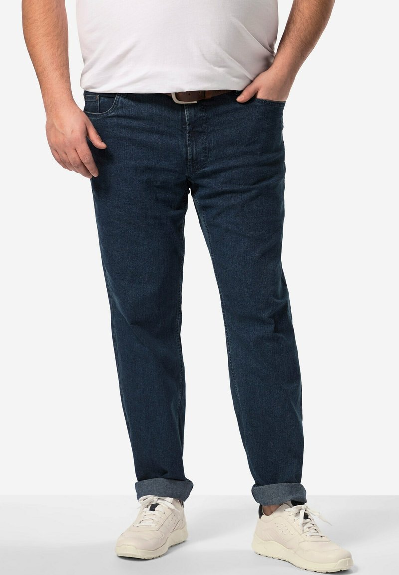 Pantalones vaqueros de mezclilla azul oscuro con un corte recto, que presentan puños doblados y bolsillos frontales. Combinados con zapatillas blancas para un look casual.
