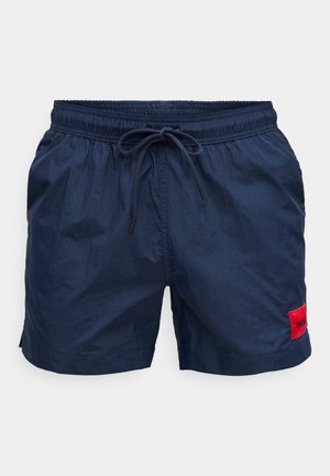 Shorts de bain bleu marine avec une ceinture élastique et un cordon de serrage, arborant un patch logo rouge sur le côté gauche et un tissu léger et texturé.