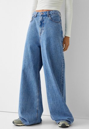 Personne portant un jean bleu taille haute à jambes larges, des baskets et un haut court blanc à manches longues, sur un fond uni.