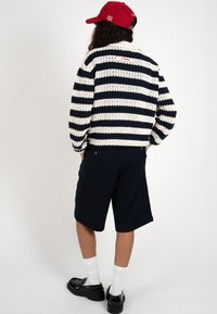 Persona con gorra roja, suéter de punto a rayas negras y blancas, pantalones cortos azules marinos, calcetines blancos y zapatos negros, de pie con las manos en los bolsillos, vista trasera.