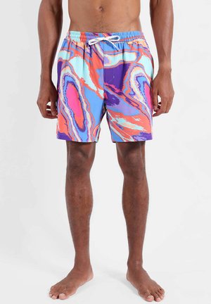 Homme portant un short de bain coloré avec des motifs abstraits violets, orange, rose et bleu, debout pieds nus sur un fond uni.