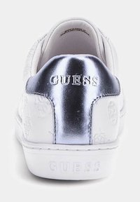 Sneaker bianco con pannello posteriore in argento lucido, motivi in rilievo sui lati e logo "GUESS" sul retro. Design a suola piatta.
