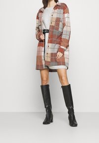 Camicia giacca oversize a quadri nei toni dell'arancione, beige e grigio, con bottoni e tasche sul petto, indossata sopra un abito grigio e stivali al ginocchio neri.