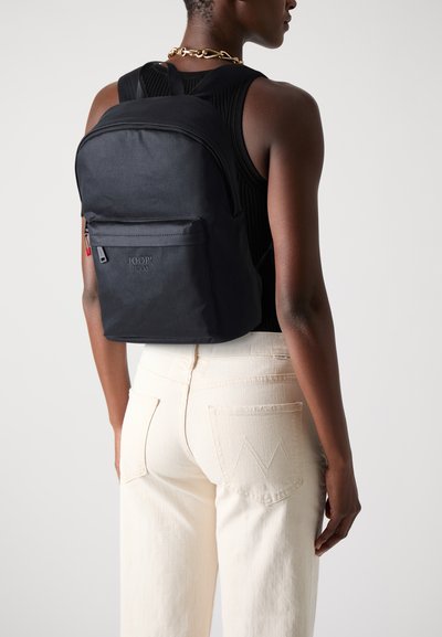 Sac à dos en nylon noir avec une poche zippée à l'avant, des bretelles ajustables et un logo embossé sur le devant. Texture lisse, design épuré.