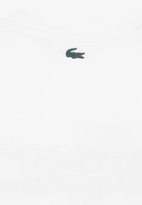 Άσπρο βαμβακερό T-shirt με κεντημένο πράσινο λογότυπο κροκόδειλου με κόκκινο στόμα στην π λάτη, ακριβώς κάτω από τη γραμμή του λαιμού.