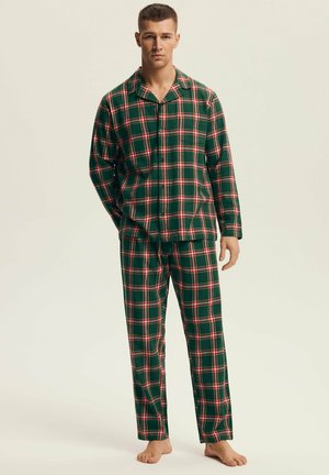 Młody mężczyzna stojący boso, ubrany w zielono-czerwono-białą kratowaną piżamę z długim rękawem i dopasowane spodnie na jednolitym tle.