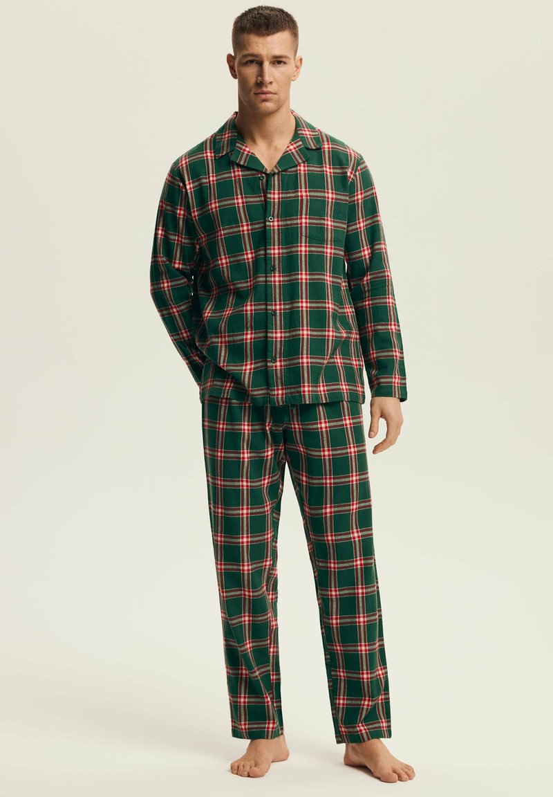 Młody mężczyzna stojący boso, ubrany w zielono-czerwono-białą kratowaną piżamę z długim rękawem i dopasowane spodnie na jednolitym tle.