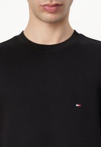 Maglione girocollo in cotone nero con un piccolo logo con dettagli rosso, bianco e blu sul lato sinistro del petto. Tessuto morbido, design aderente.
